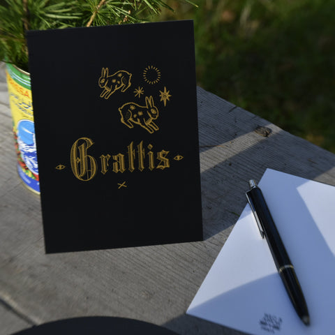 A black greeting card with yellow rabbit illustrations and the word 'Grattis' (Swedish for Congratulations) stands on a weathered wooden table next to a white card backside, a black pen, and a can with a plant outdoors.